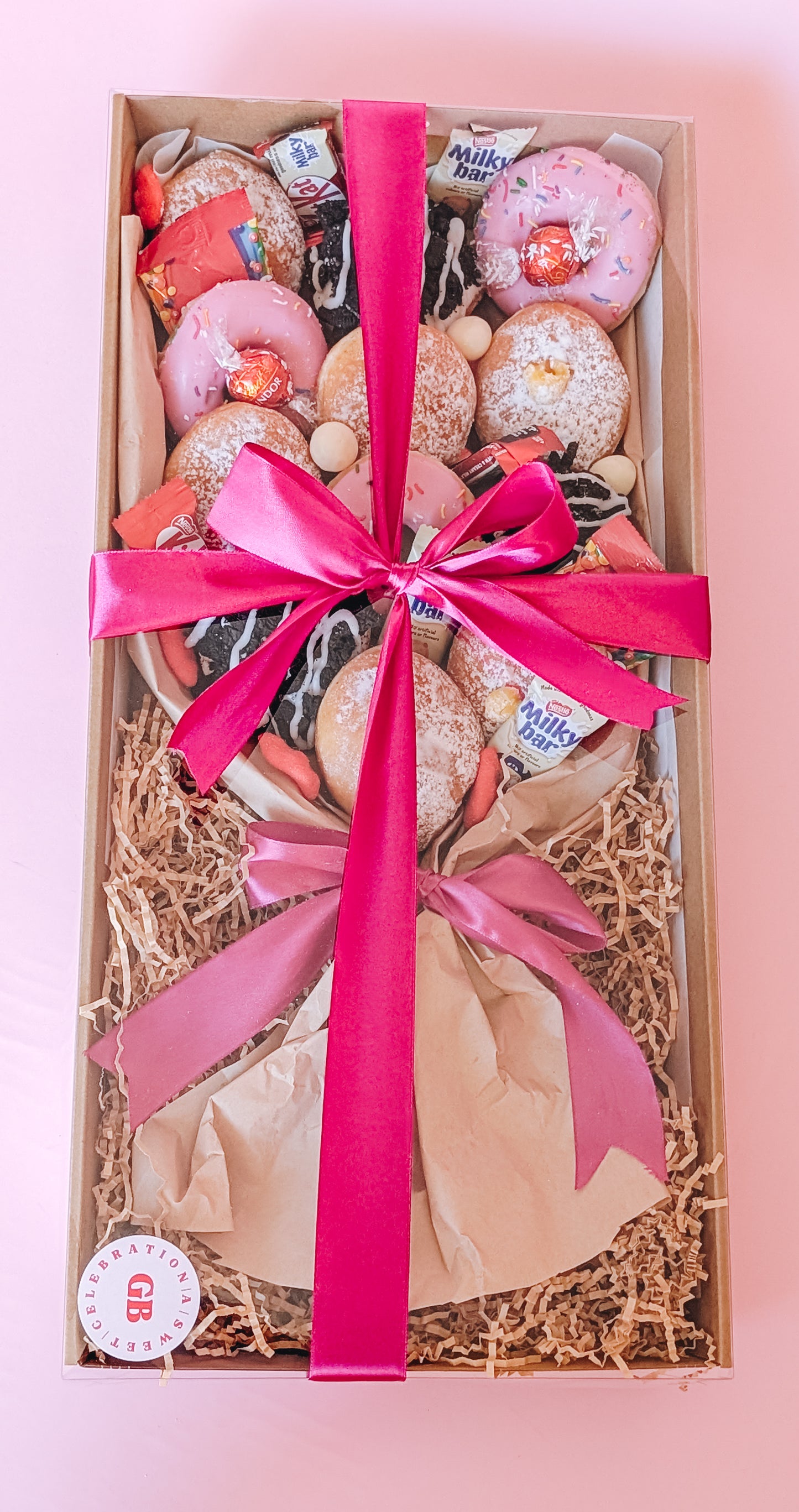 Photograph of a box of donuts arranged in a bouquet style. The box also contains an assortment of fun size chocolates and lollies. The box is wrapped in a bright pink silk ribbon tied in a bow.