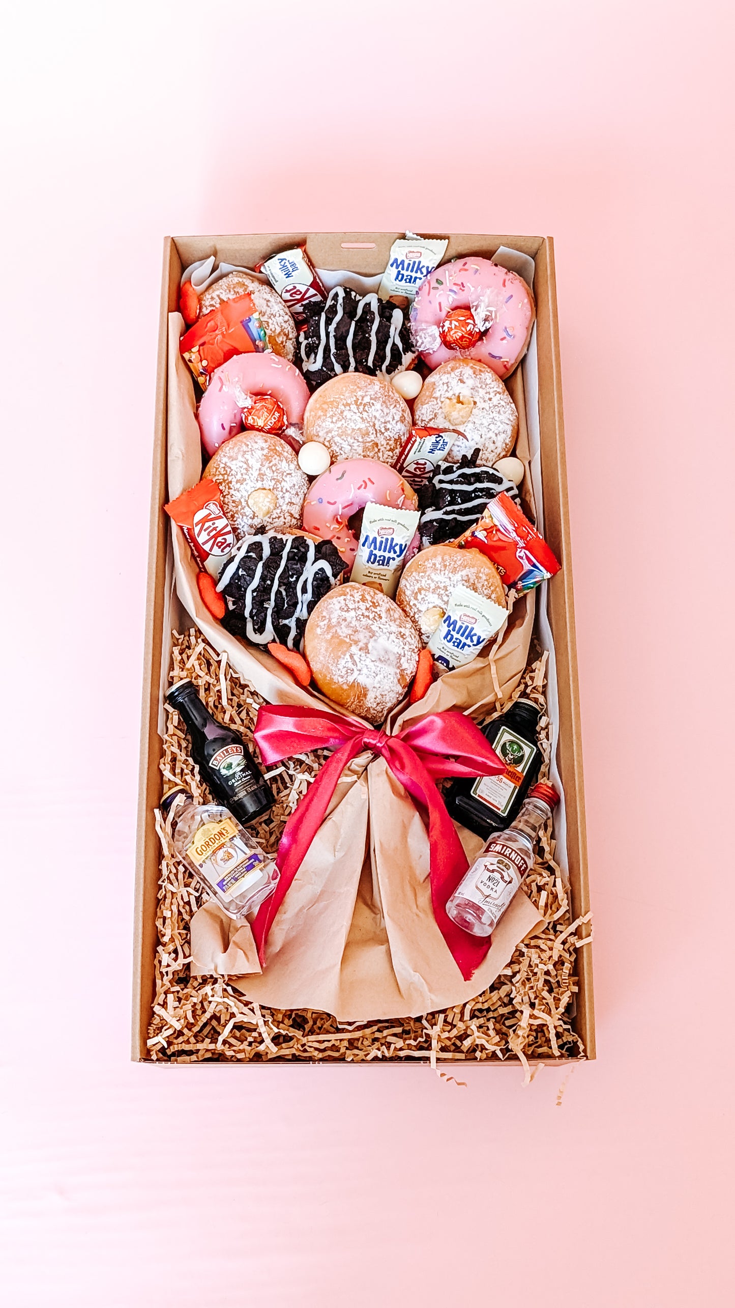 Photograph of a dessert box donut bouquet far away. It is filled with twelve donuts that are different flavours including pink icing sprinkles, chocolate oreo, lemon curd and jam. The box has mini chocolates and sweets along with 4 x miniature alcohols.