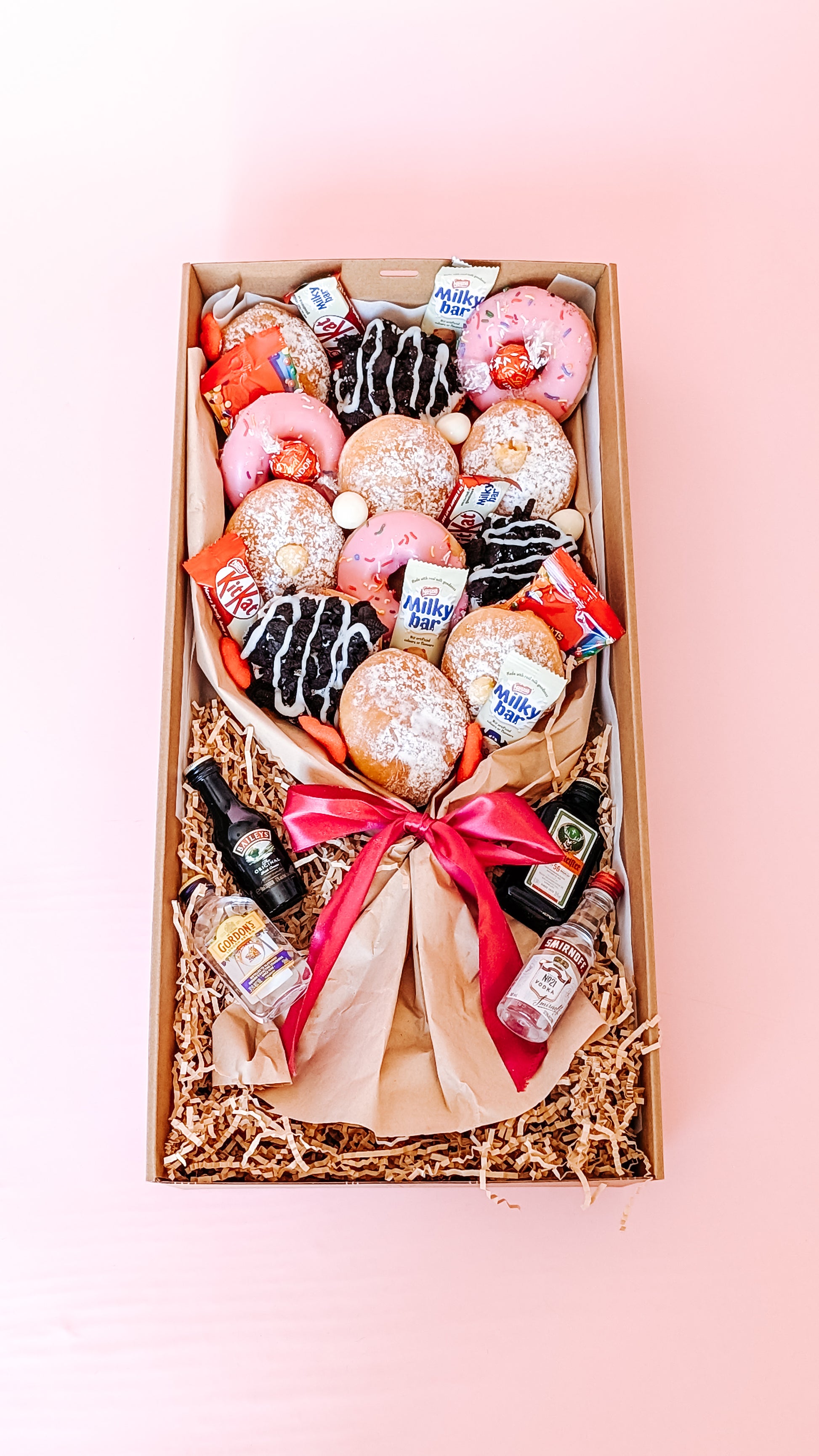 Photograph of a dessert box donut bouquet far away. It is filled with twelve donuts that are different flavours including pink icing sprinkles, chocolate oreo, lemon curd and jam. The box has mini chocolates and sweets along with 4 x miniature alcohols.