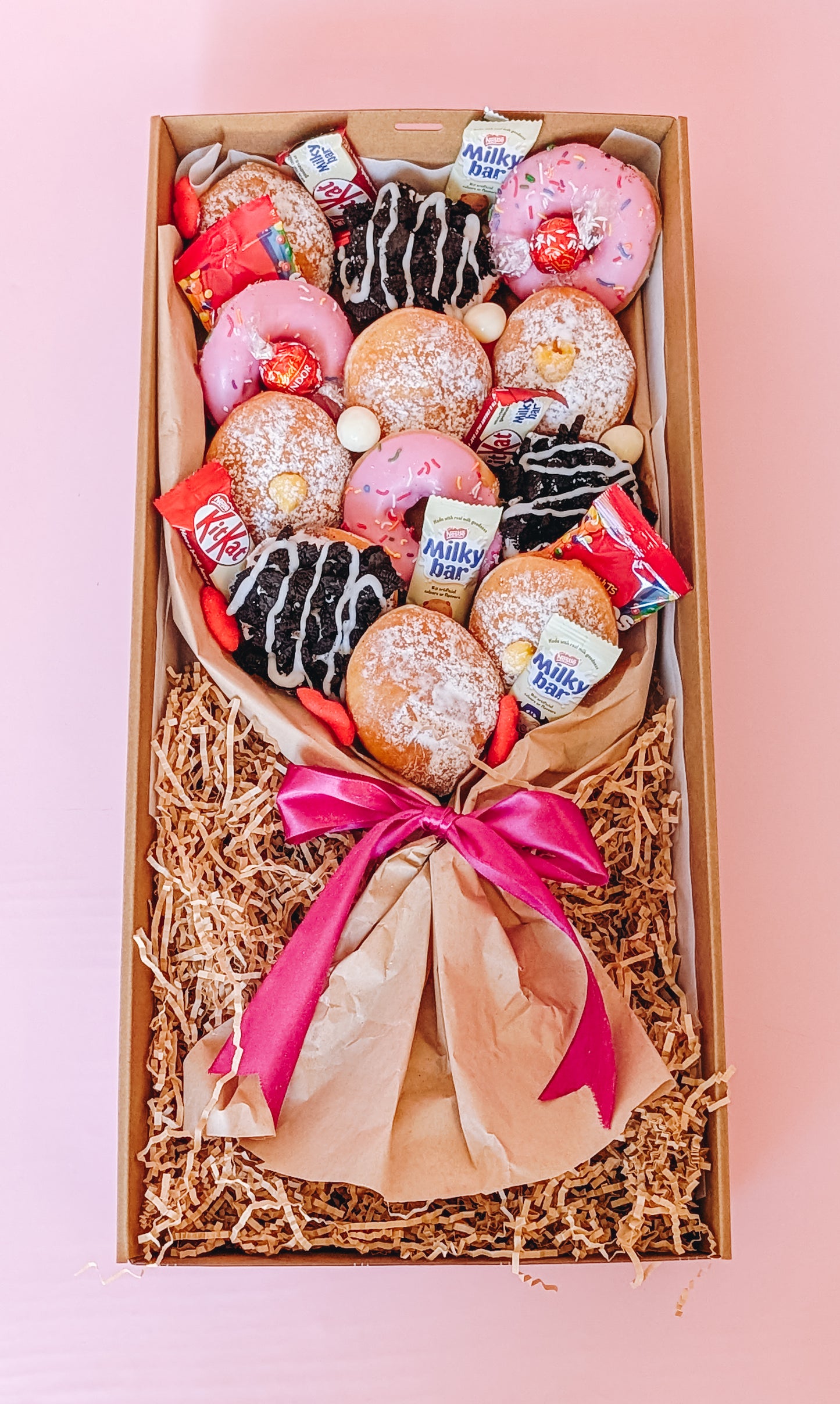 Photograph of a box of donuts arranged in a bouquet style. The box also contains an assortment of fun size chocolates and lollies in an ariel view.