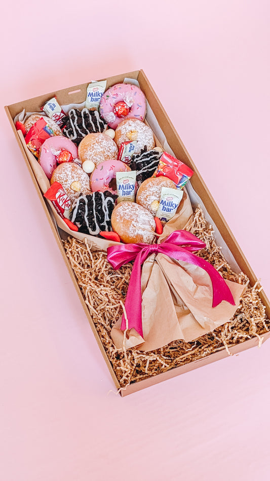 Photograph of a box of donuts arranged in a bouquet style. The box also contains an assortment of fun size chocolates and lollies.