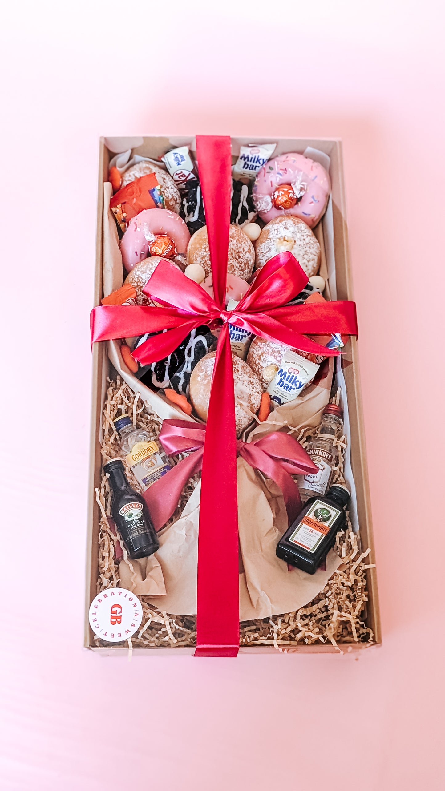 Photograph of a dessert box donut bouquet. It is filled with twelve donuts that are different flavours including pink icing sprinkles, chocolate oreo, lemon curd and jam. The box has mini chocolates and sweets along with 4 x miniature alcohols. The box is wrapped in a silk ribbon pink bow.