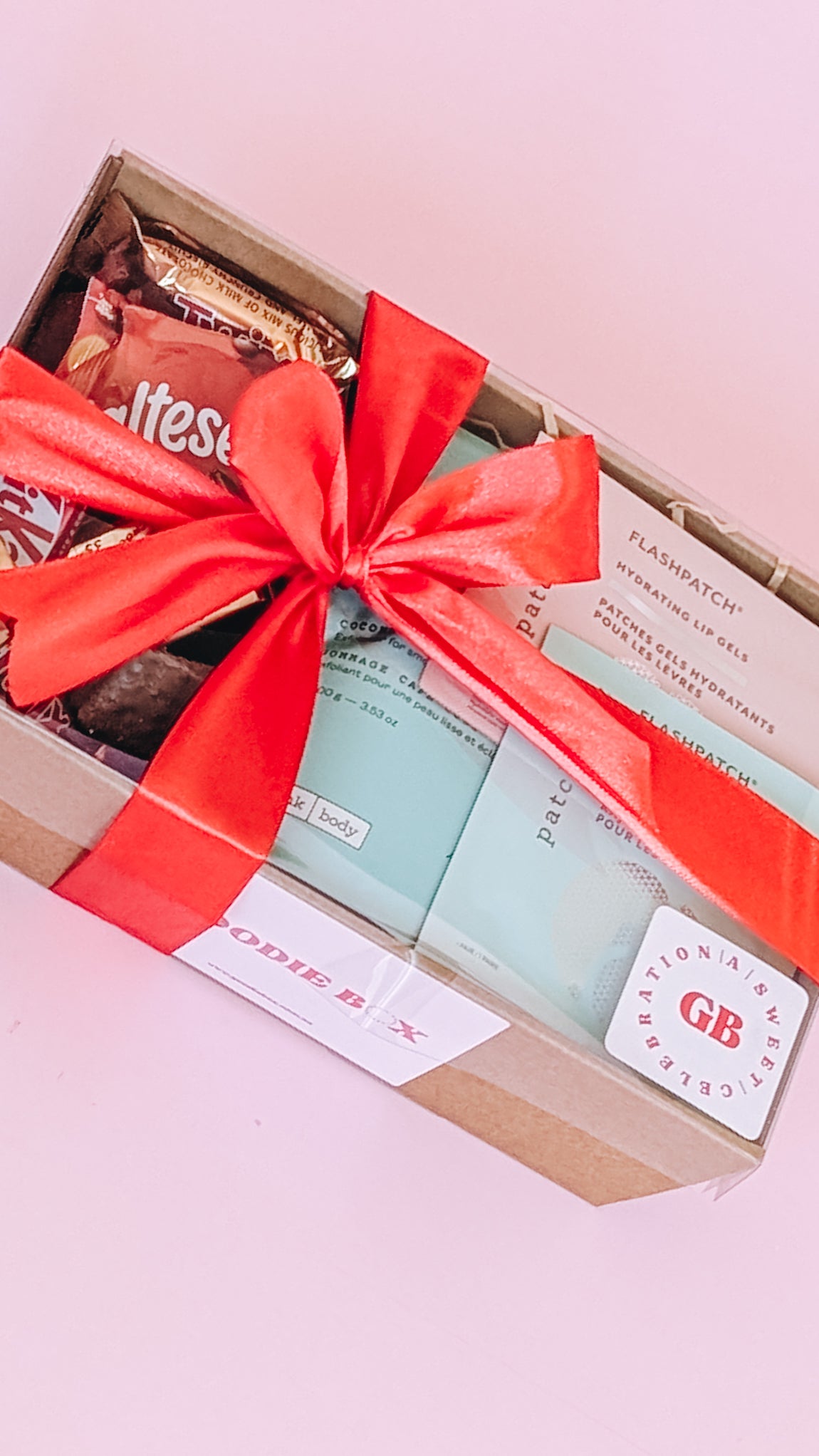 Photograph of pamper hamper wrapped in a red silk bow. The box is filled with pampering goods such as Frank Body scrub, eye mask and lip mask and contains an assortment of sweet treats.