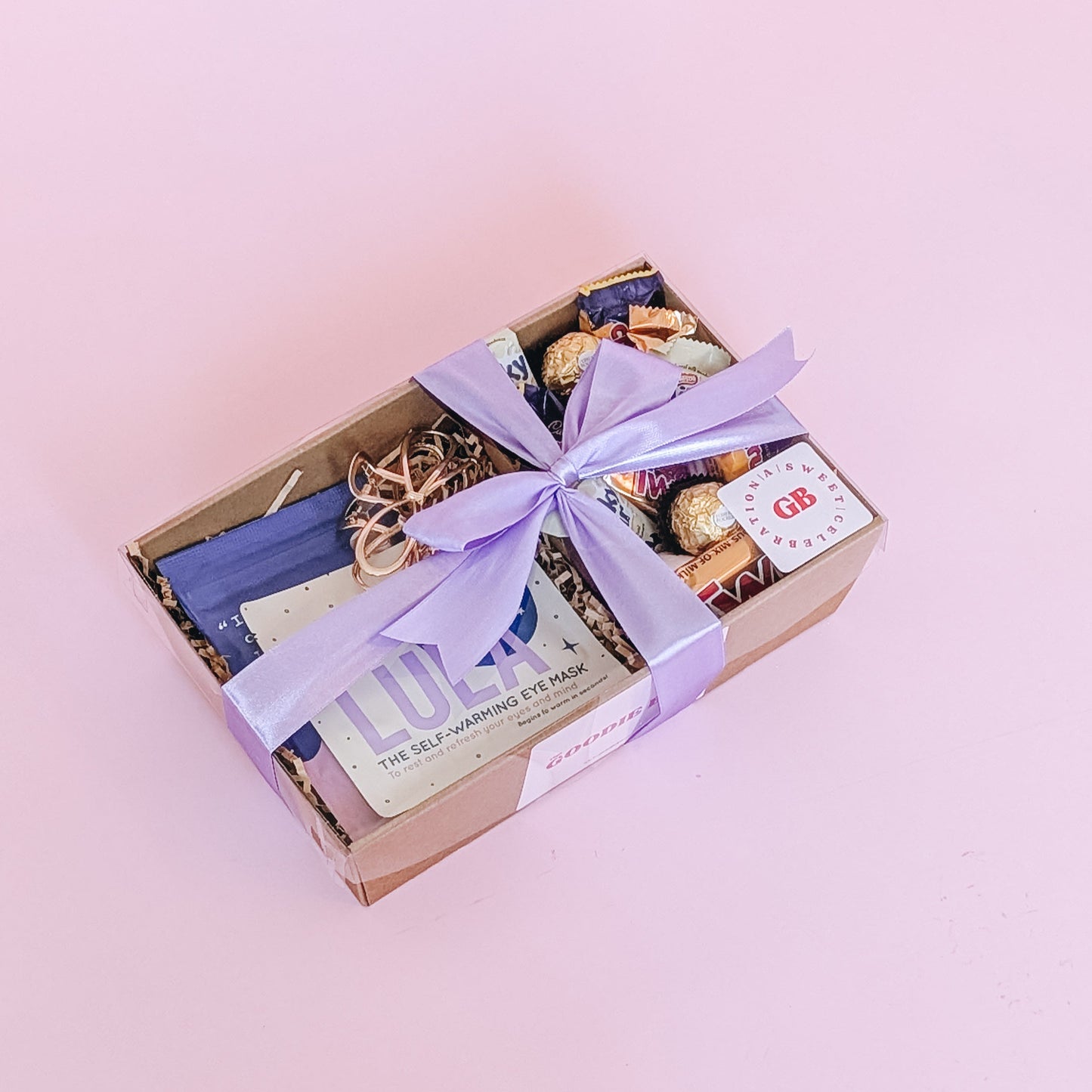 Photograph of pamper hamper sleepy time filled with a selection of sweet treats, including marshmallows, Crunchie, Milky Bar, Twix & Ferrero Rocher. There are self care items in the gift hamper including a gold hair claw, frank body scrub, and Lula self warming eye mask. The gift hamper is wrapped with a lilac silk ribbon bow.