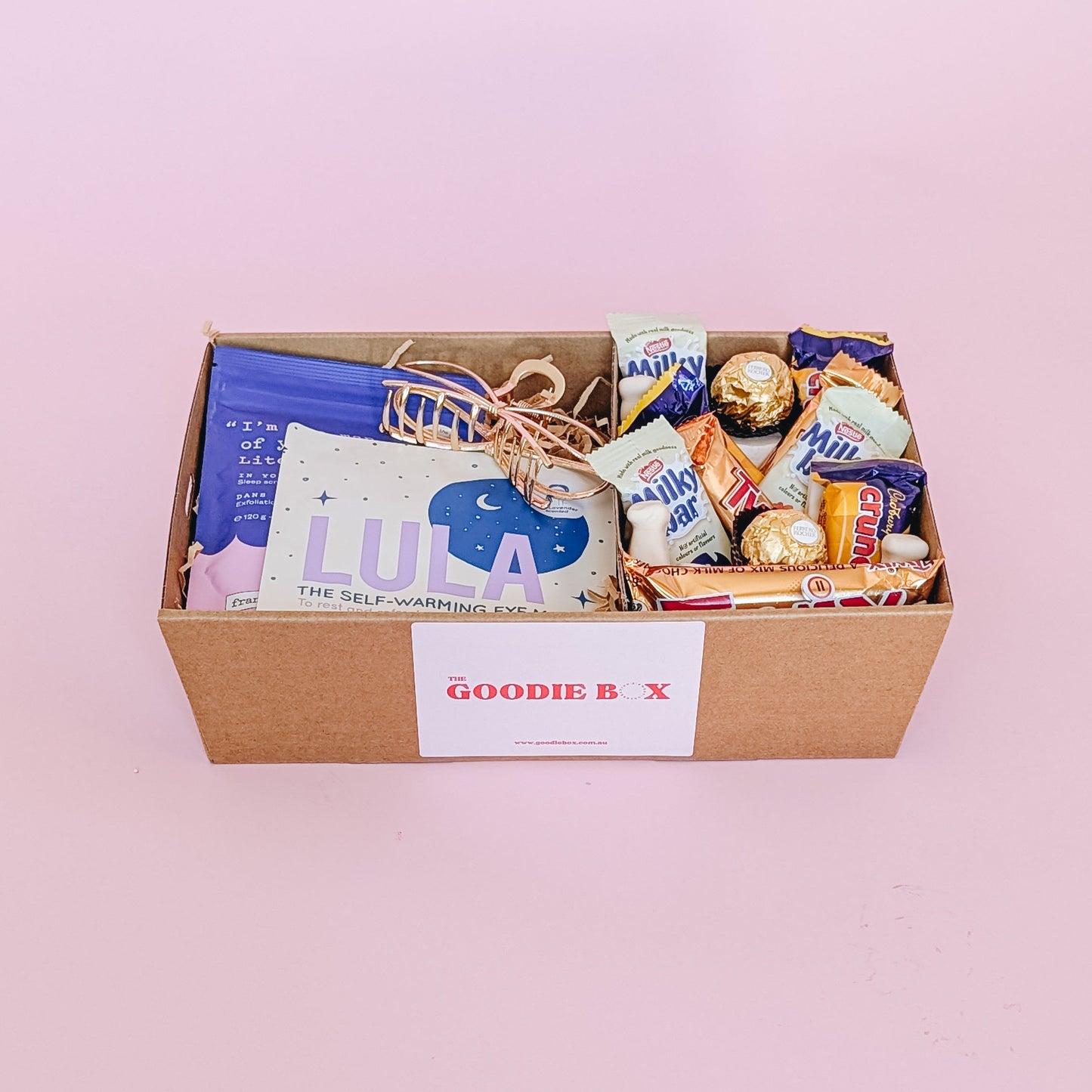 Photograph of pamper hamper sleepy time filled with a selection of sweet treats, including marshmallows, Crunchie, Milky Bar, Twix & Ferrero Rocher. There are self care items in the gift hamper including a gold hair claw, frank body scrub, and Lula self warming eye mask.