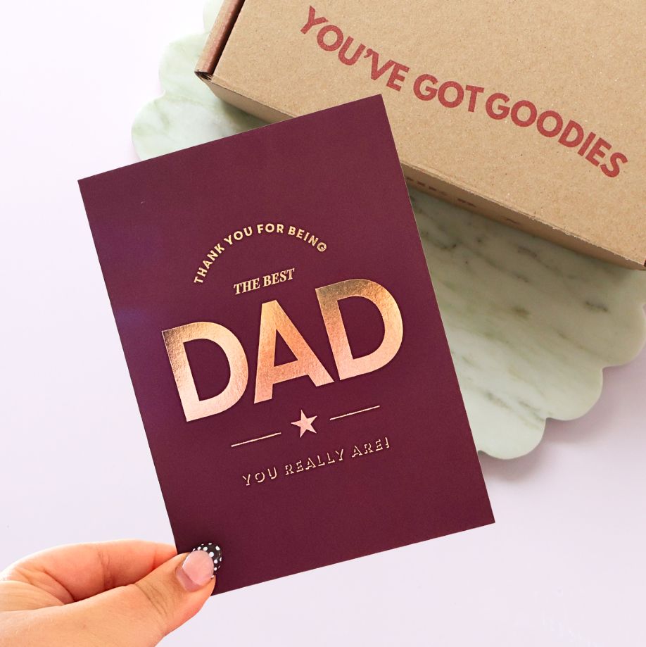 A hand holds a burgundy card with metallic gold foil text reading 'THANK YOU FOR BEING THE BEST DAD YOU REALLY ARE!' in front of a brown gift box printed with 'YOU'VE GOT GOODIES' on a light background.