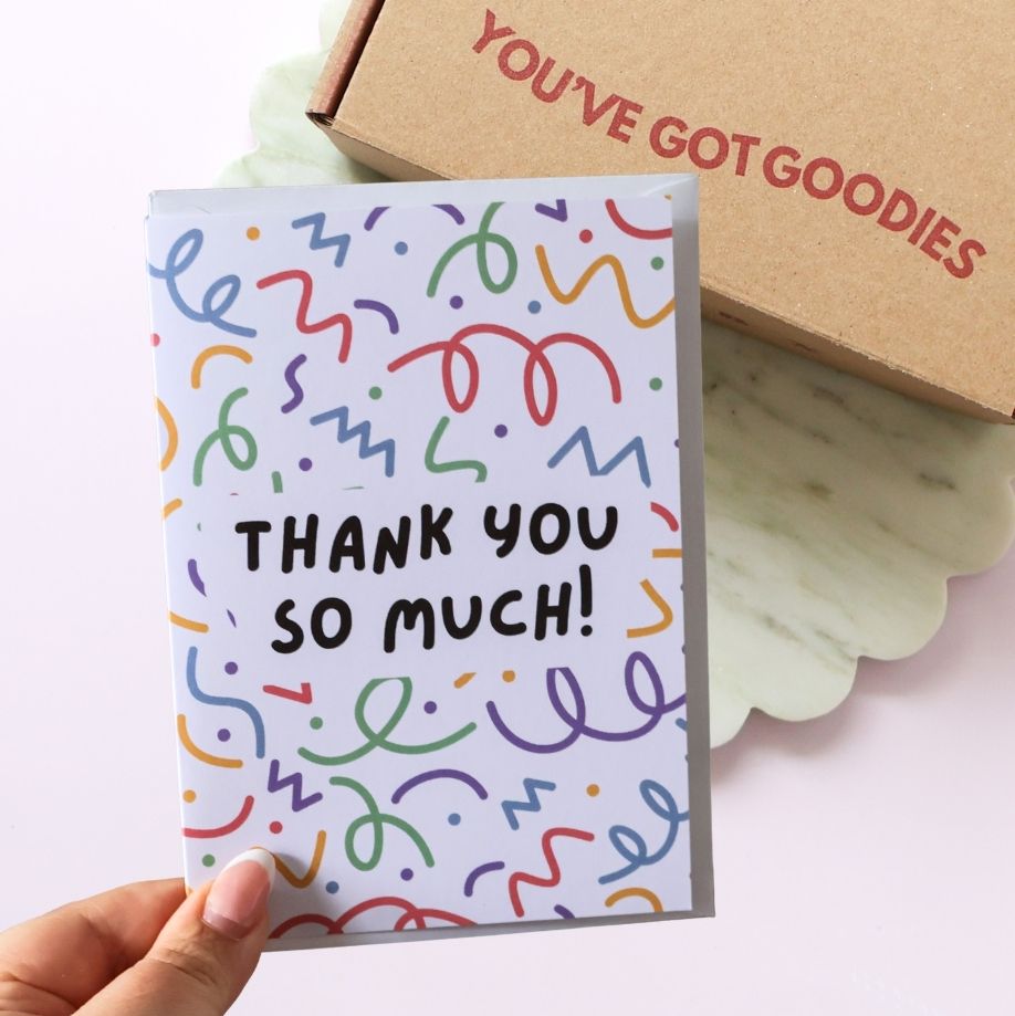 Card with colorful text 'THANK YOU SO MUCH!' held by a hand, with a box labeled 'YOU'VE GOT GOODIES' in the background.