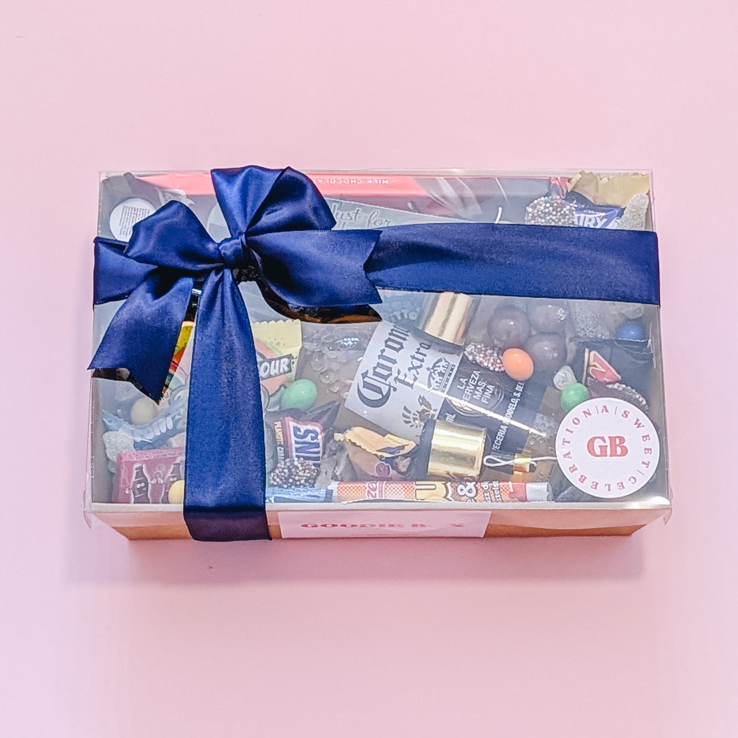 Photograph of gift box filled with Corona, Nerds, Cadbury Dairy Milk, Trolli Lollies and an assortment of mini chocolates and loose cookies and sweets. Wrapped in a silk blue ribbon bow and a can of cider.