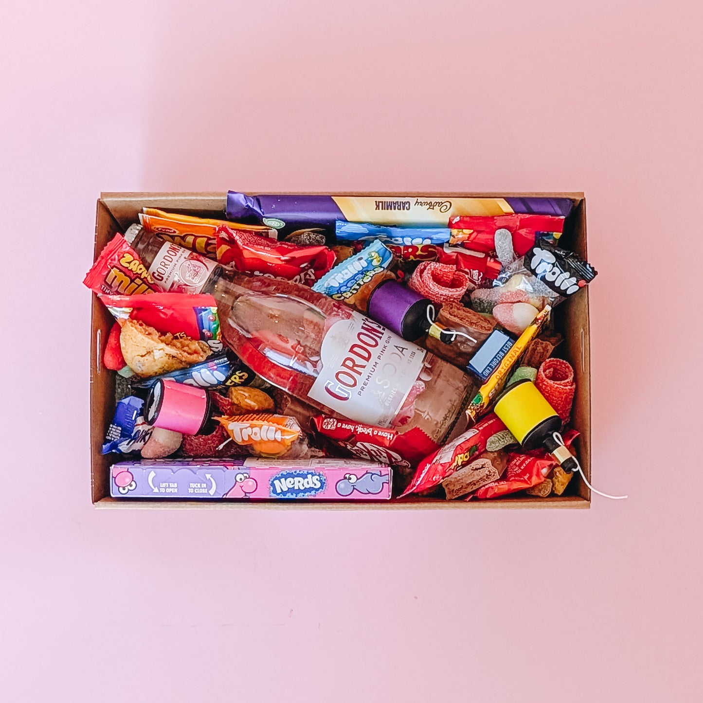 Photograph of gift box filled with Gordon's pink gin, Nerds, Cadbury CaraMilk, Trolli Lollies and an assortment of mini chocolates and loose cookies and sweets.