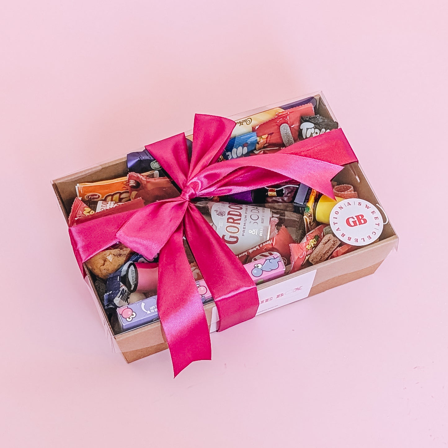 Photograph of gift box filled with Gordon's pink gin, Nerds, Cadbury CaraMilk, Trolli Lollies and an assortment of mini chocolates and loose cookies and sweets. wrapped in a bright pink silk ribbon bow.