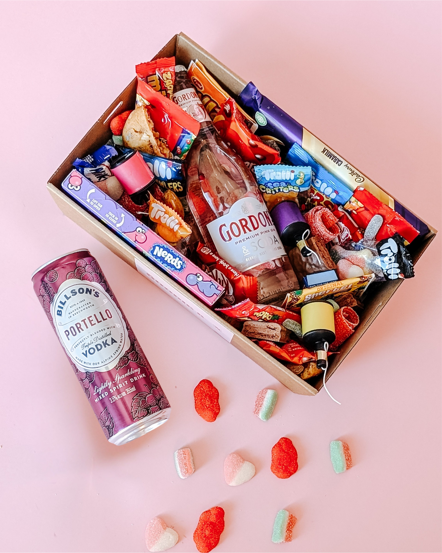 Photograph of gift box filled with Gordon's pink gin, Nerds, Cadbury CaraMilk, Trolli Lollies and an assortment of mini chocolates and loose cookies and sweets. Loose sweets and Billson's vodka.