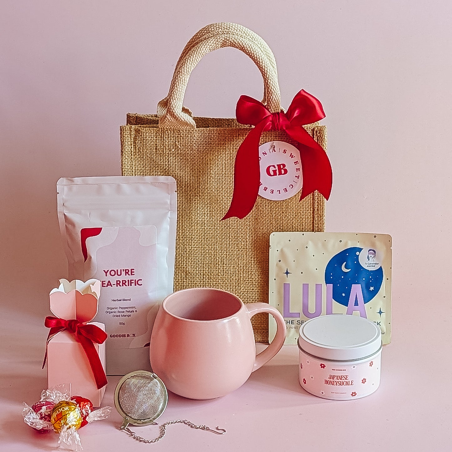 Jute tote bag with herbal tea, lula eye mask, candle, pink mug and lindt truffles on a pink background.