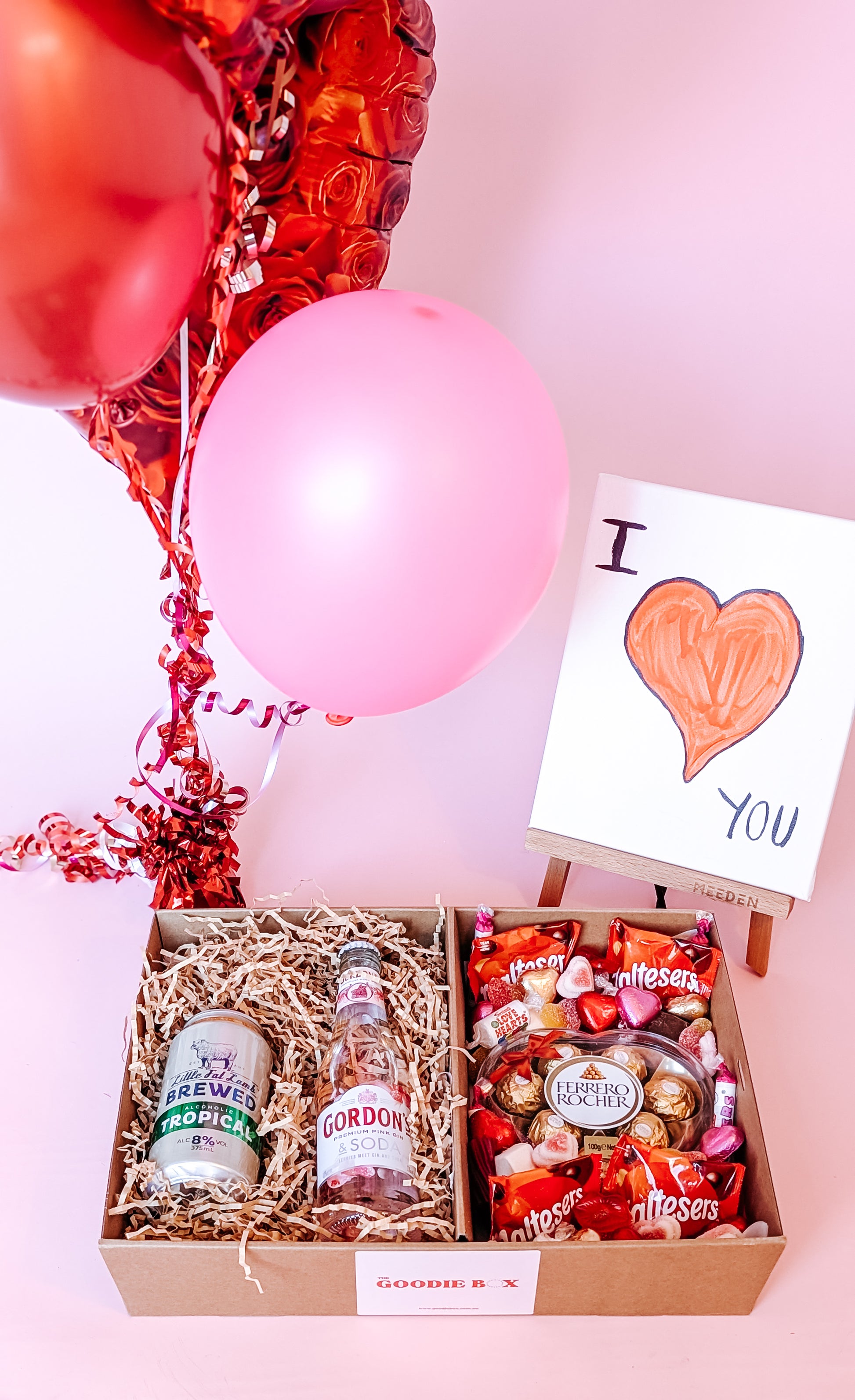 photograph of valenitne's romantic gift hamper. It includes a can of cider, a pink gin, and an assortment of chocolates and lollies including: a ferrero Rocher heart, maltesers, swizzels love hearts and love themed sweets. There is pink & red balloons in the background and an easel with a canvas painted with "I Love You".