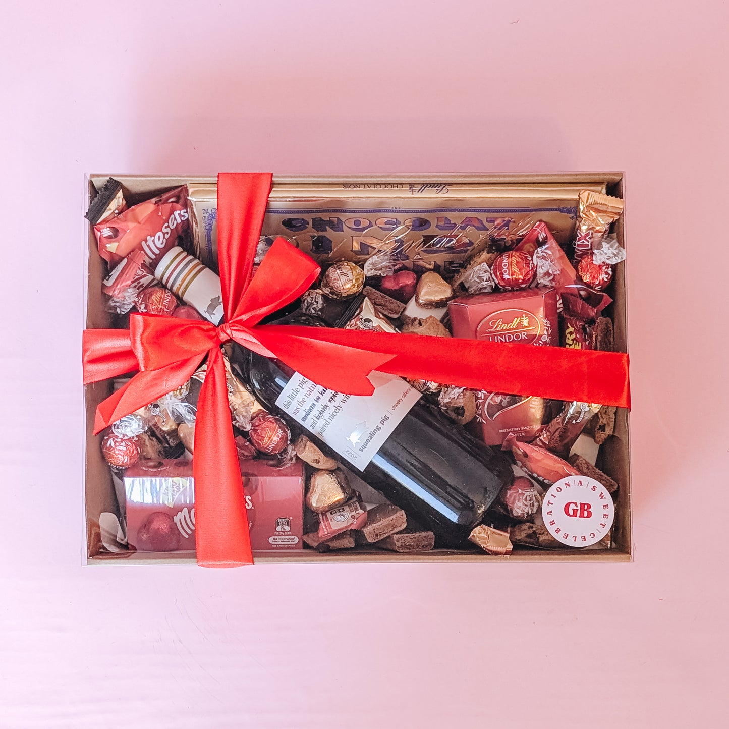 Photograph of indulgent valentines gift hamper. The box is filled with romantic themed chocolates and lollies and a bottle of red wine. Wrapped in a silk red ribbon bow.