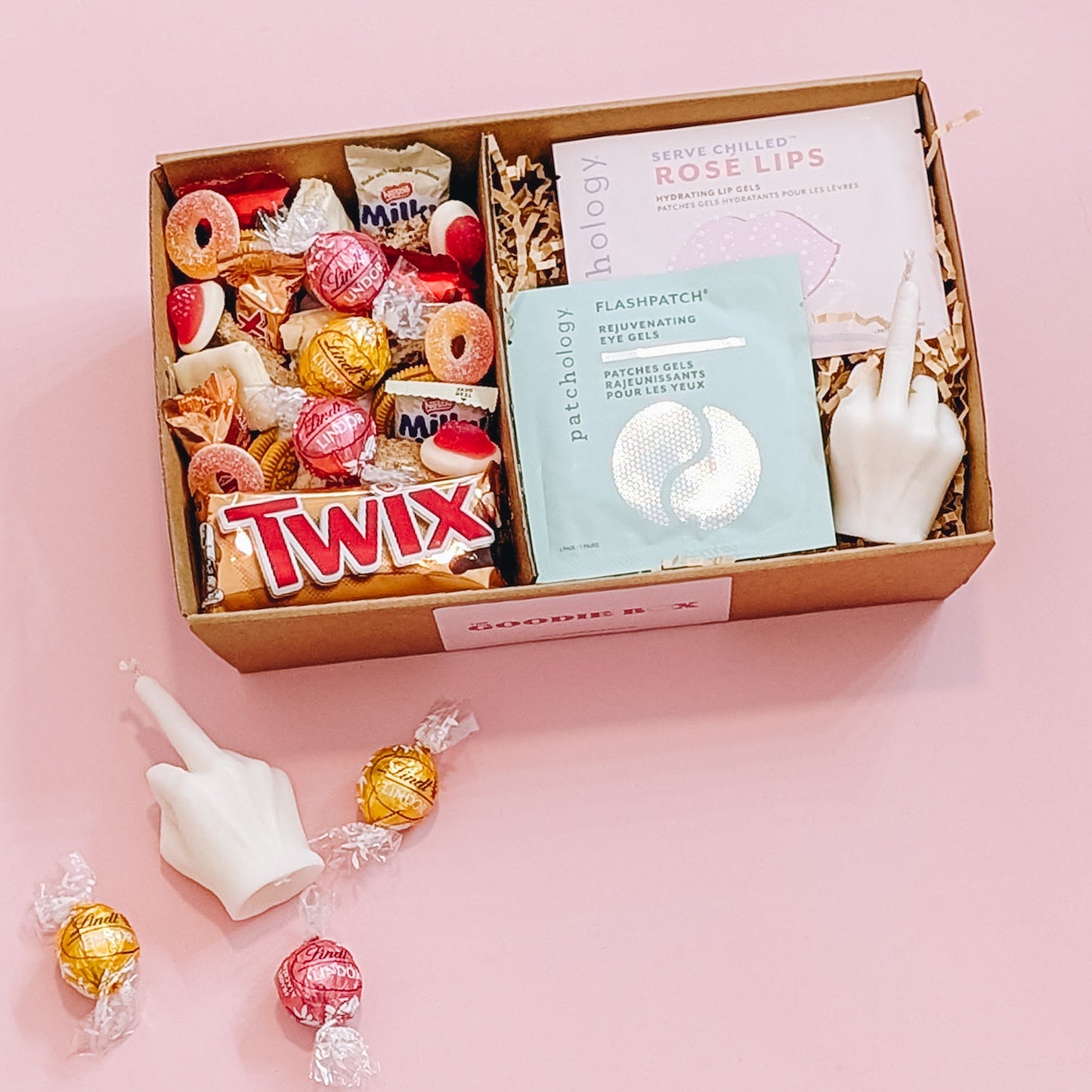 Photograph of the better off without him gift pamper hamper. The gift box is filled with a patchology eye and lip mask, a rude finger candle and an assortment of gold and pink chocolate, lollies, and cookies. There are marshmallows, chocolate, candle scattered on the floor.