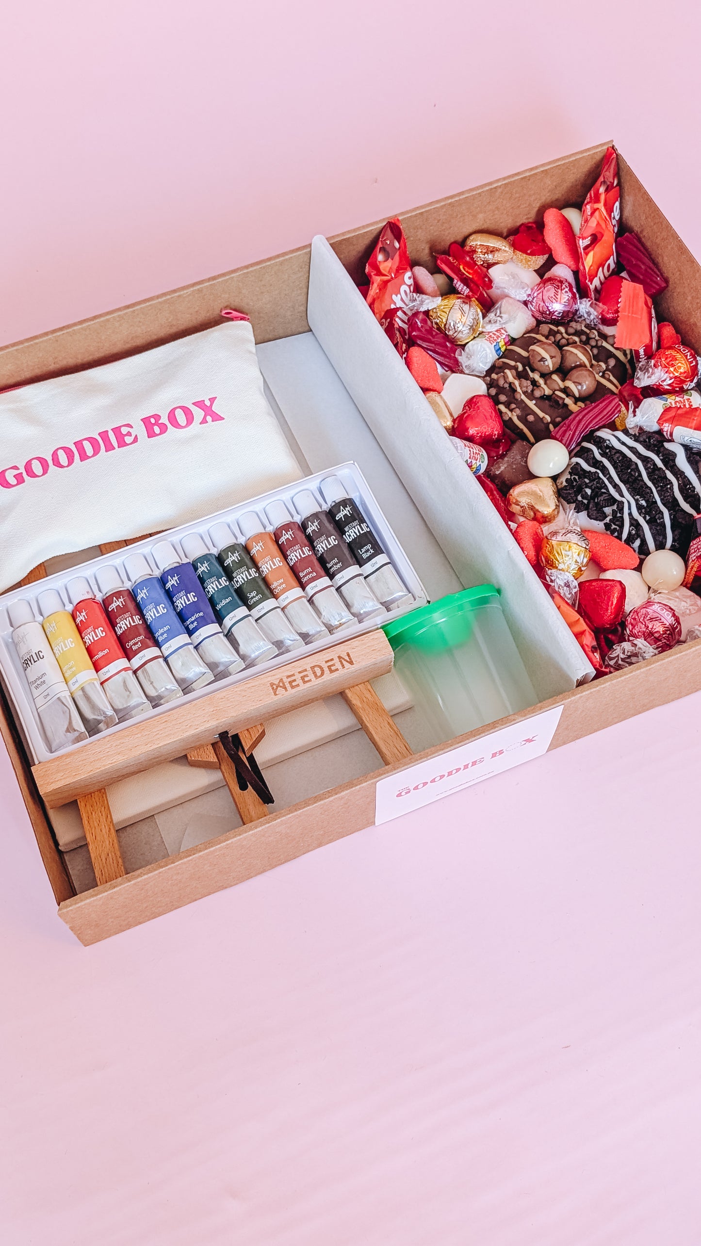 Photograph of the date night in a box - nice edition. The left hand side of the box is filled with art easels, canvases, an acrylic paint set, and a pencil case. The other half is filled with two donuts and an assortment of chocolates and lollies that are red themed for valentines day.