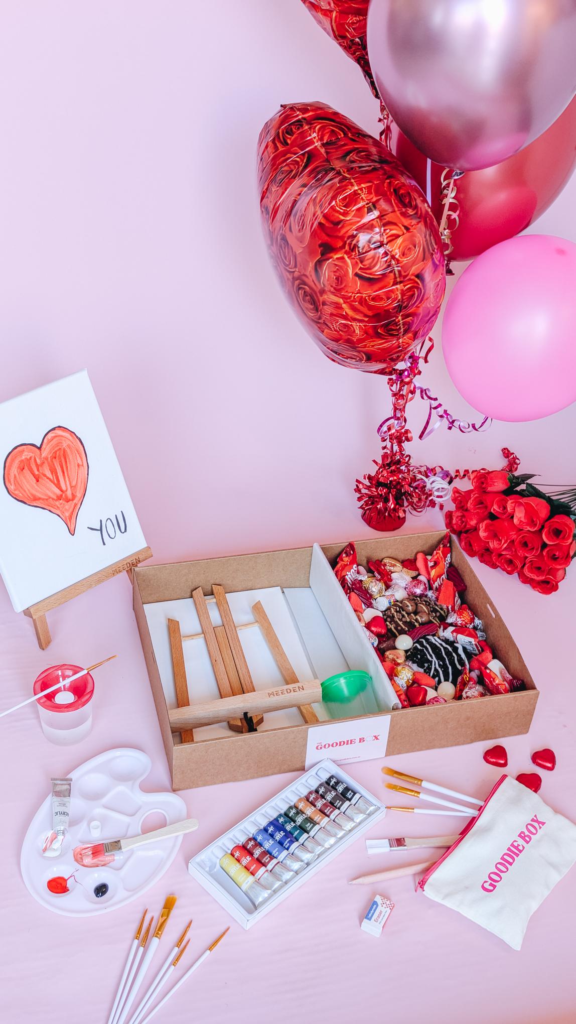Photograph of the date night in a gift box - nice edition. The left hand side of the box is filled with art easels, canvases, an acrylic paint set, and a pencil case. The other half is filled with two donuts and an assortment of chocolates and lollies that are red themed for valentines day.