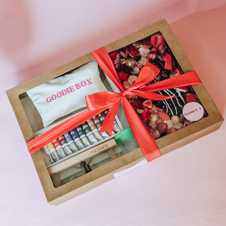 Photograph of the date night in a dessert box - nice edition. The left hand side of the box is filled with art easels, canvases, an acrylic paint set, and a pencil case. The other half is filled with two donuts and an assortment of chocolates and lollies that are red themed for valentines day. Wrapped in a red silk ribbon tied in a bow.