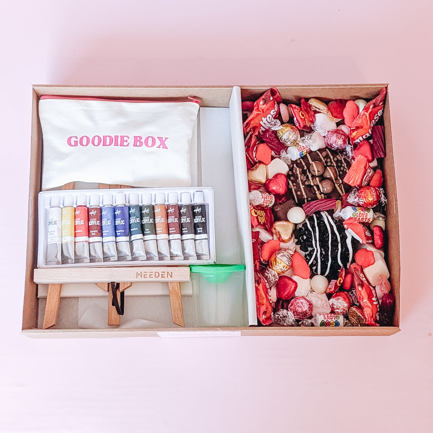 Photograph of the date night in a box - nice edition. The left hand side of the box is filled with art easels, canvases, an acrylic paint set, and a pencil case. The other half is filled with two donuts and an assortment of chocolates and lollies that are red themed for valentines day.