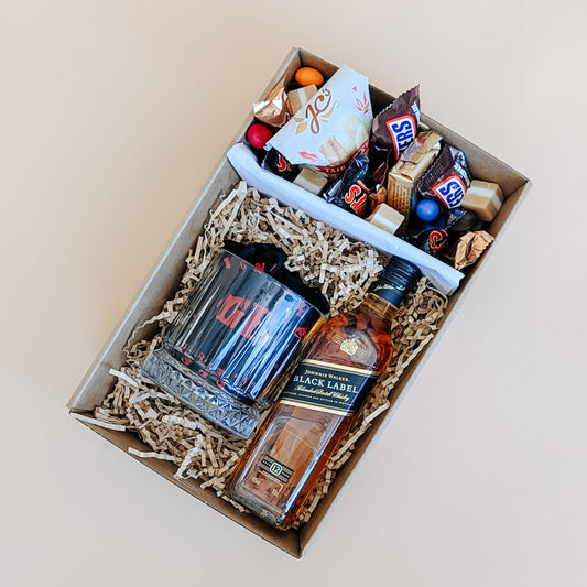 Photograph of whisky gift set for him. The gift hamper is filled with a bottle of Johnny Walker whisky, a whisky glass and a ice cube. The other half is filled with chocolates, nuts and fudge.