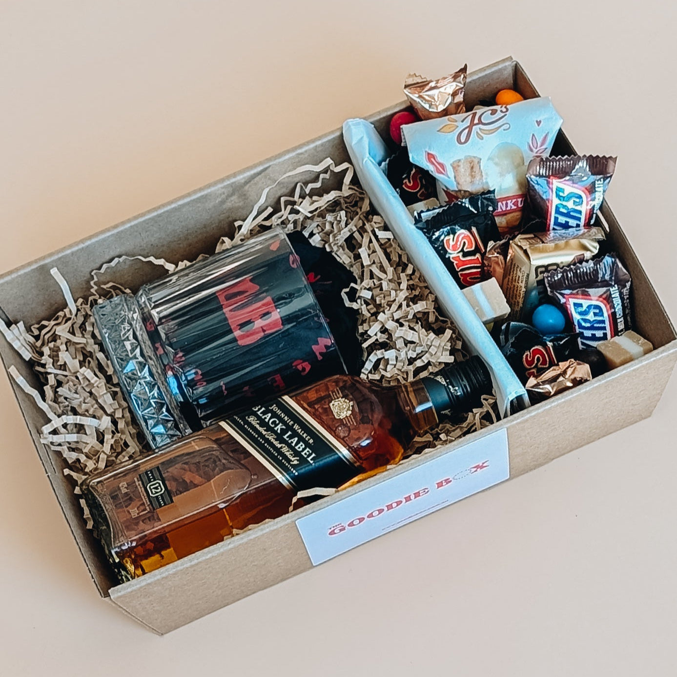 Photograph of whisky gift set for him. The gift hamper is filled with a bottle of Johnny Walker whisky, a whisky glass and a ice cube. The other half is filled with chocolates, nuts and fudge.