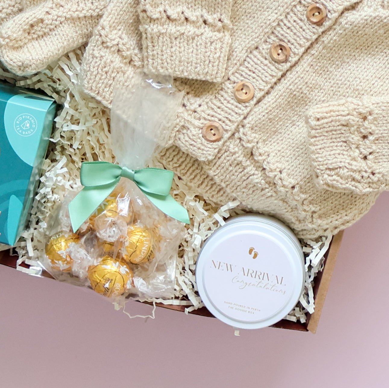 Baby gift basket with knitted cardigan, chocolate, and baby items on a pink background