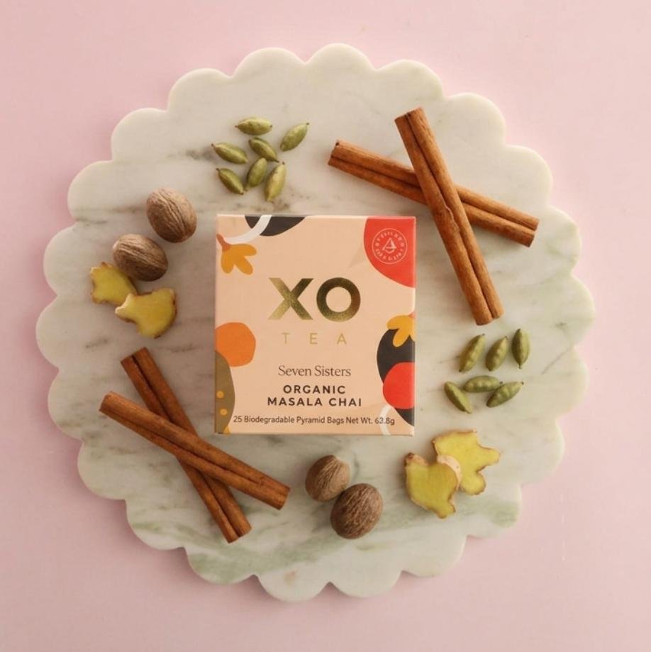 XO Tea Seven Sisters Organic Masala Chai in a peach-toned box, surrounded by cinnamon sticks, cardamom pods, whole nutmeg and fresh ginger on a scalloped marble board.