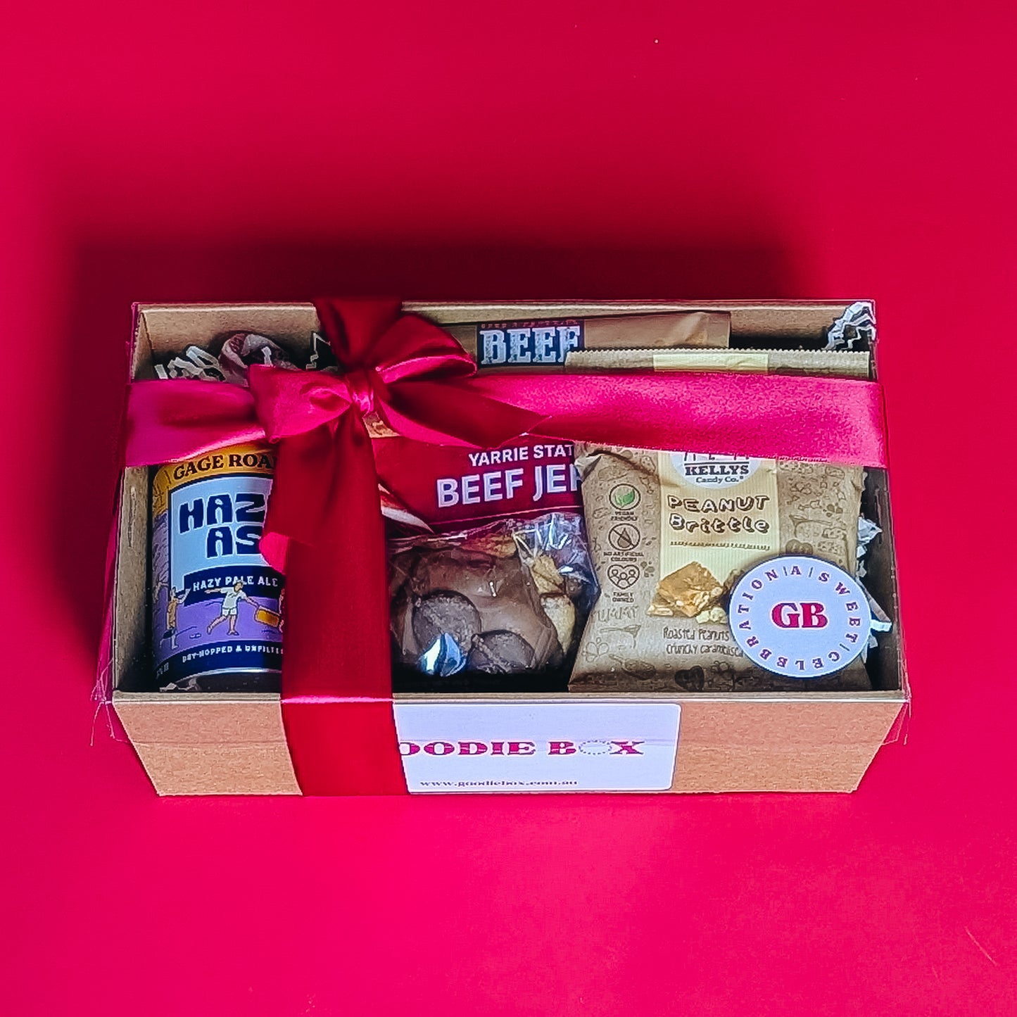 corporate gift box with beef jerkie, pale ale peanut brittle and cookie with a red ribbon on a red background.