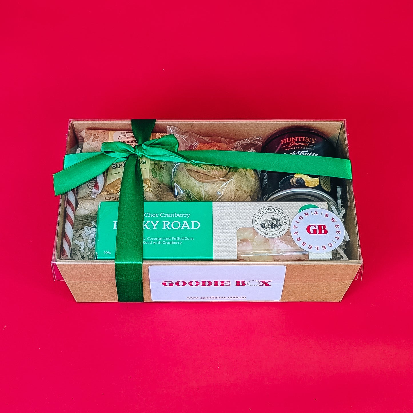 Gift box with rocky road, cookie, chips and peanut brittle with a green bow on a red background.