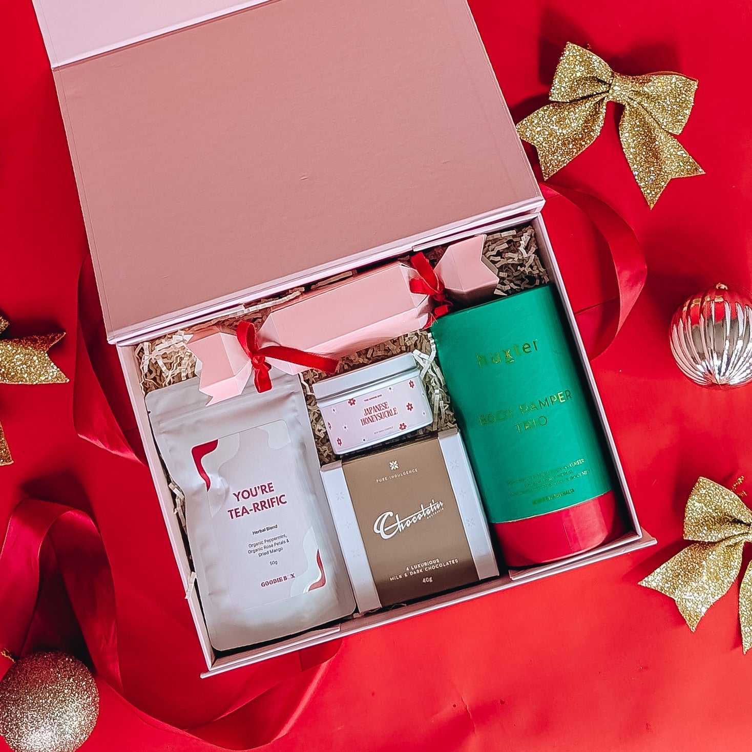 Gift hamper filled with chocolate, bath and body goods, herbal tea and a candle on a red background