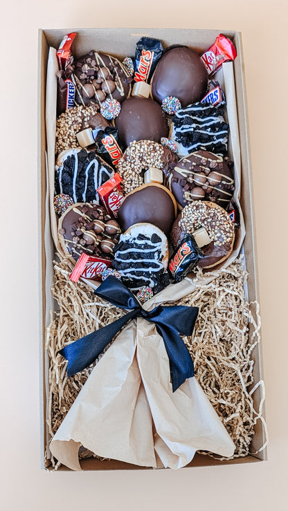 Photograph of a box of donuts arranged in a bouquet style. The box also contains an assortment of fun size chocolates and lollies.