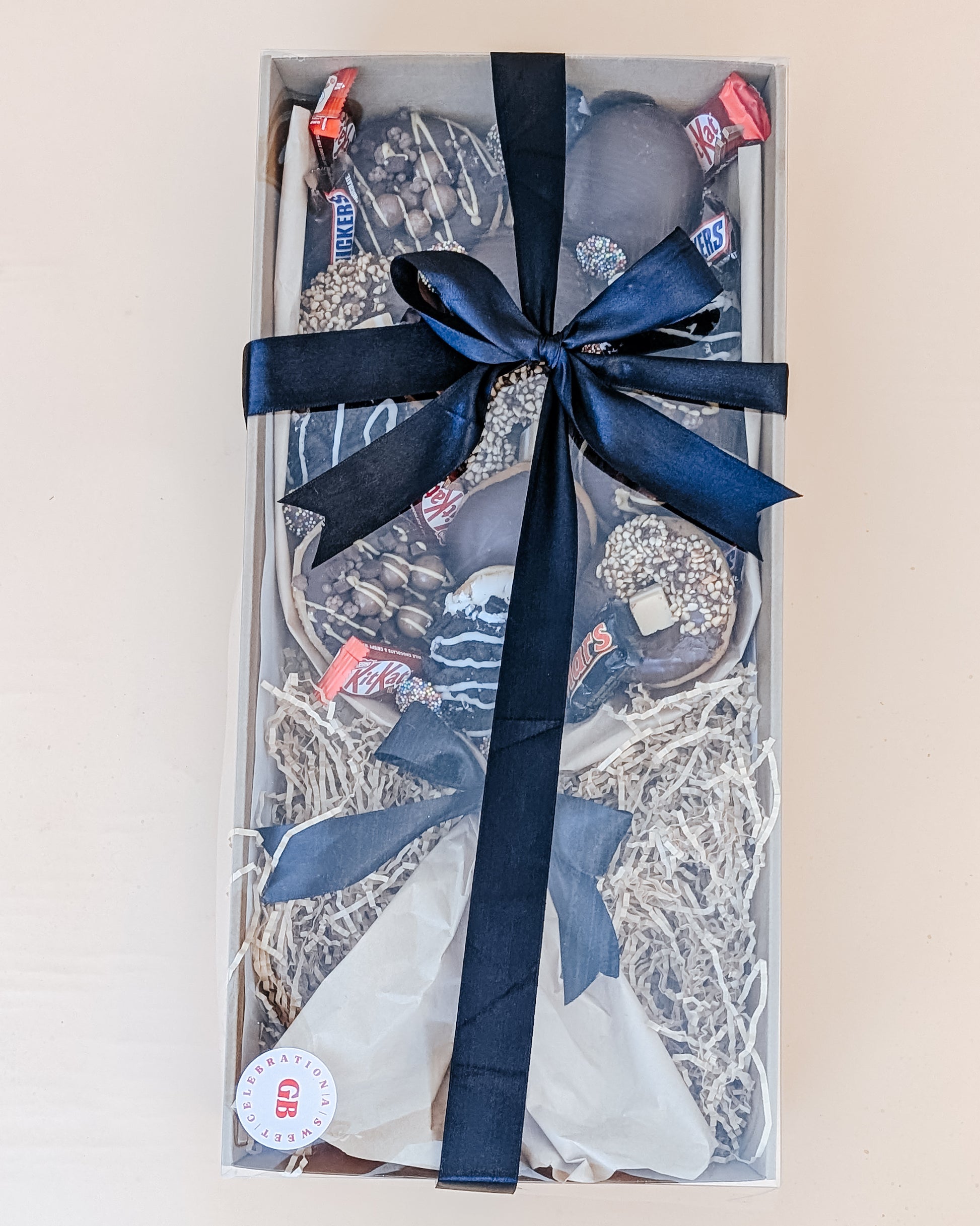 Photograph of a box of donuts arranged in a bouquet style. The box also contains an assortment of fun size chocolates and lollies. Tied in a black silk ribbon bow.