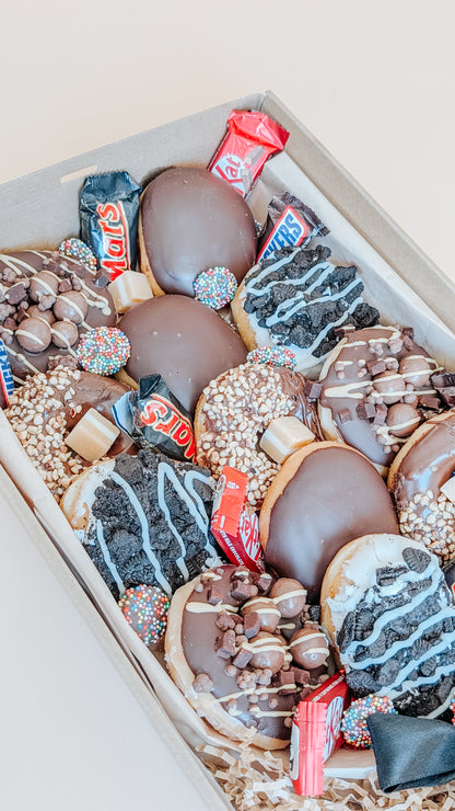 Photograph of a box of donuts arranged in a bouquet style close up. The box also contains an assortment of fun size chocolates and lollies.