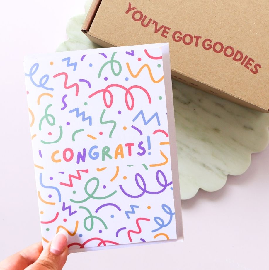 Colorful 'CONGRATS!' card held by a hand with a brown box labeled 'YOU'VE GOT GOODIES' in the background.