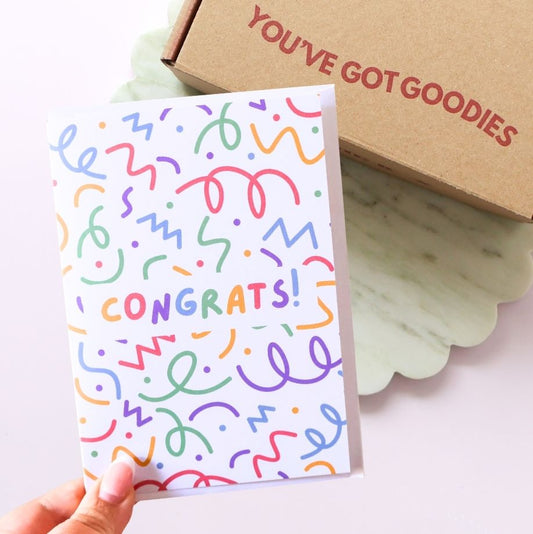 Colorful 'CONGRATS!' card held by a hand with a brown box labeled 'YOU'VE GOT GOODIES' in the background.