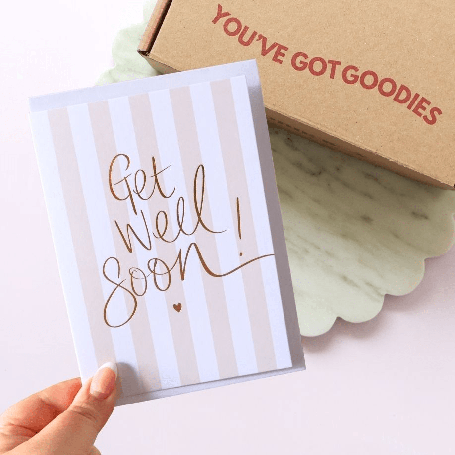 Hand holding a 'Get Well Soon' card with a box labeled 'You've Got Goodies' in the background.