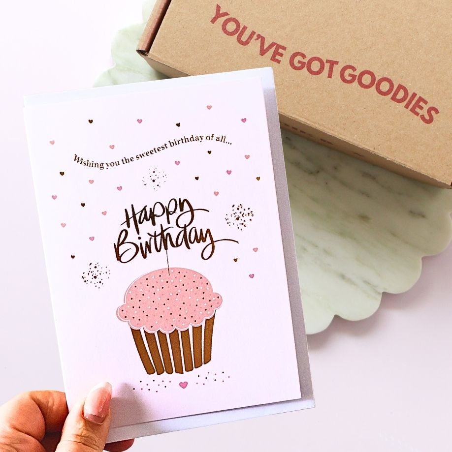 Birthday card with pink cupcake illustration and 'Happy Birthday' text, held by a hand with a box labeled 'YOU'VE GOT GOODIES' in the background.
