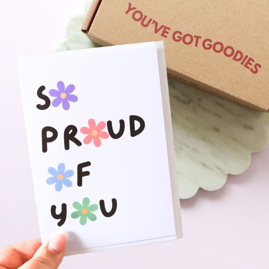 Card with 'S PROUD F YOU' text and flowers, held by a hand, with a box labeled 'YOU'VE GOT GOODIES' in the background.