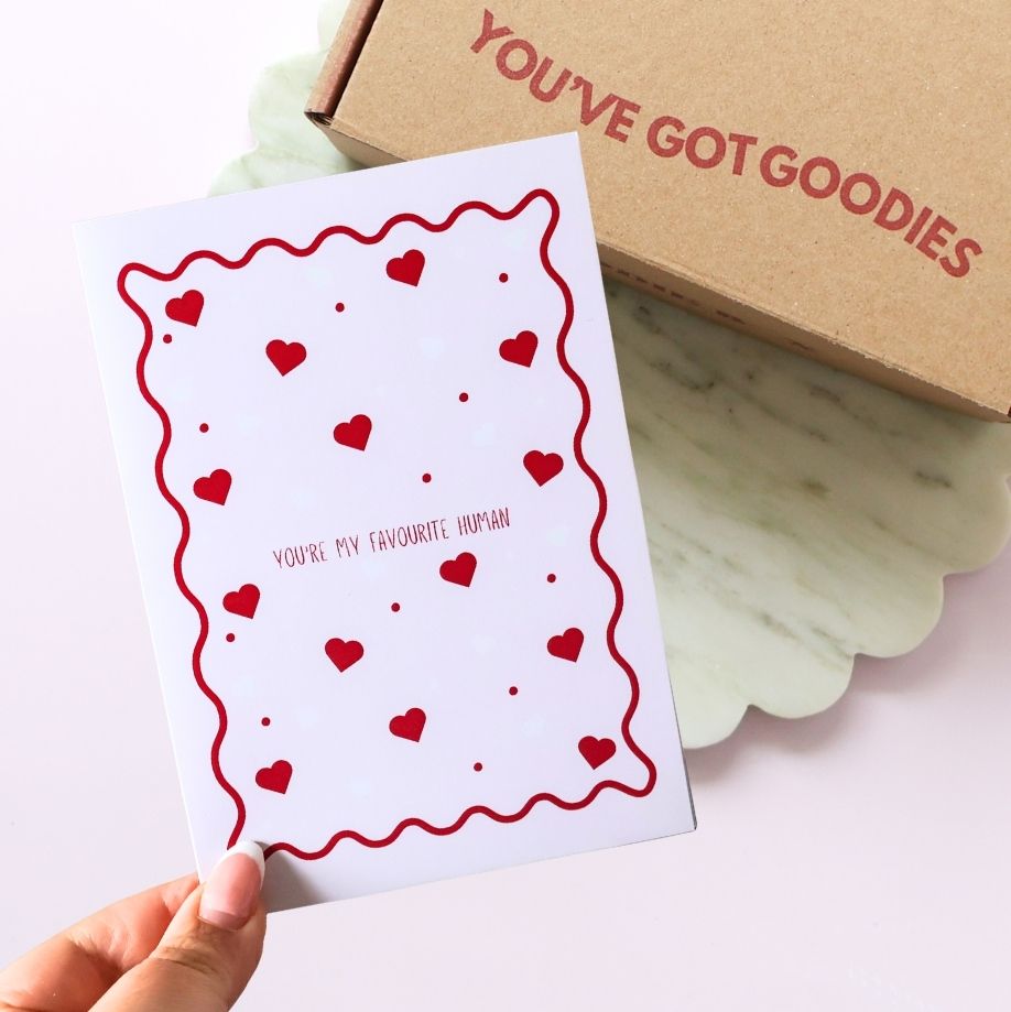 Card with red heart pattern and 'You're my favourite human' text, held by a hand, with a box labeled 'You've Got Goodies' in the background.
