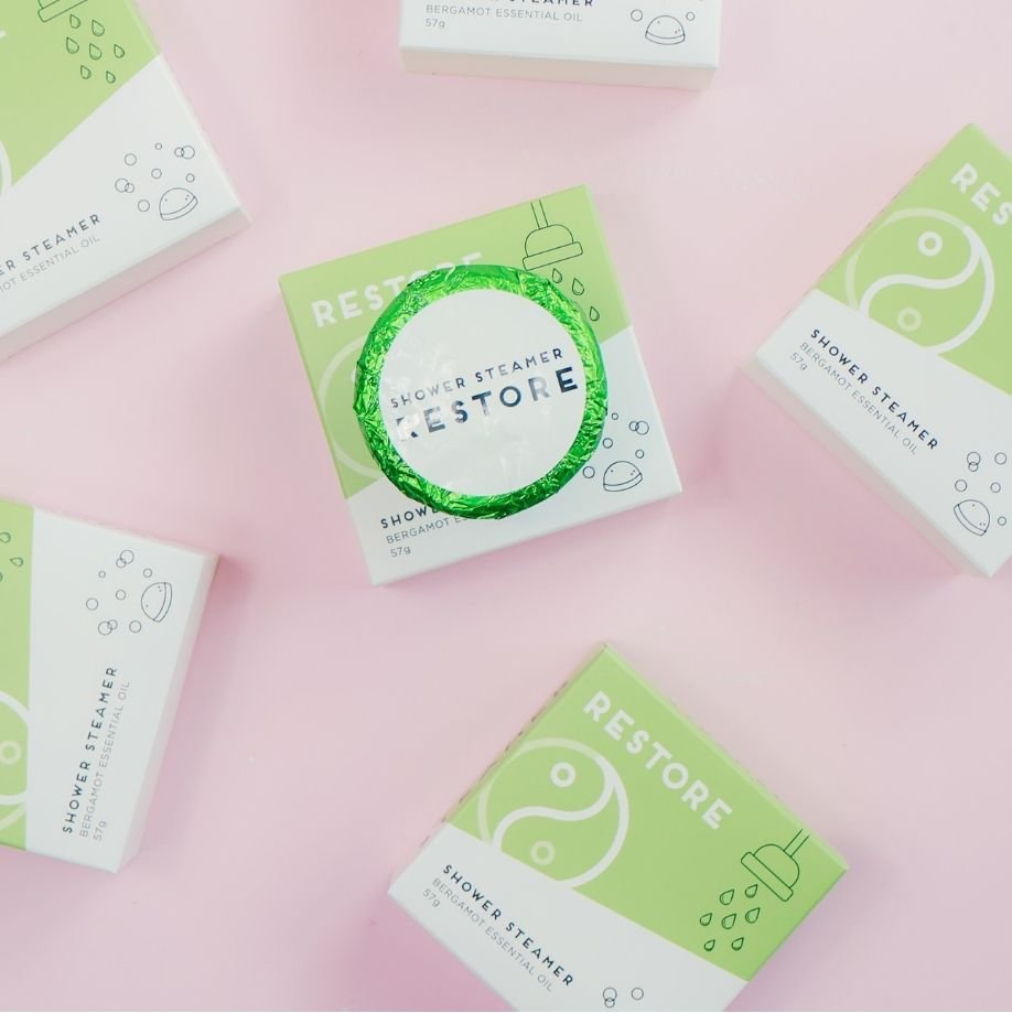 Green and white 'Restore' bergamot shower steamer boxes on a pink background