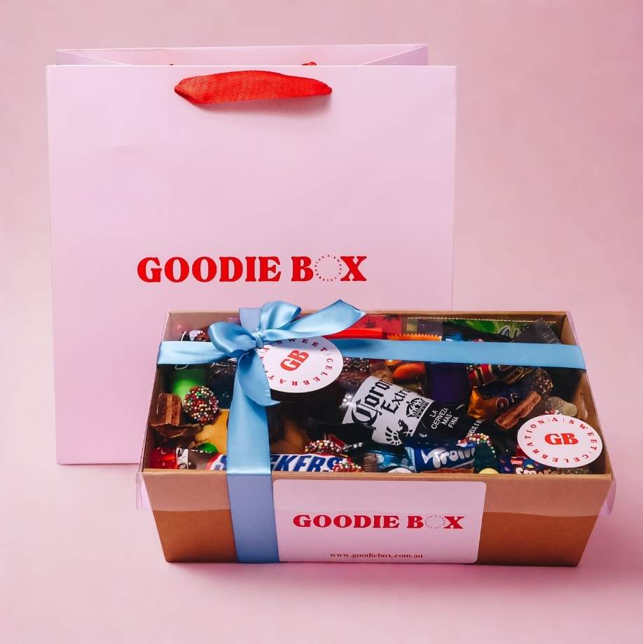 Goodie Box birthday gift hamper for him with Corona beer, Snickers and assorted lollies in branded kraft box with blue ribbon and Goodie Box gift bag on pink background