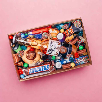 Overhead view of Goodie Box dessert hamper for him with Corona Extra beer, Snickers, Mars, Smarties, Trolli gummies, loaded cookie and assorted lollies on pink background