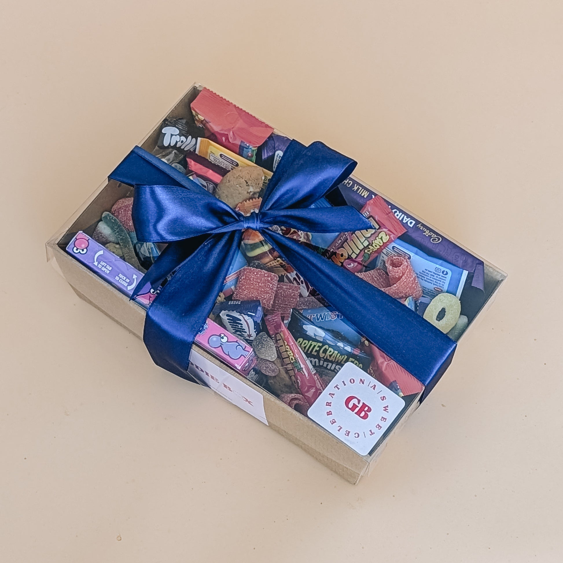 Photograph of birthday dessert gift box. It is filled with chocolate bars, a selection of lollies, cookies and party poppers. The box is wrapped with a blue silk ribbon bow.