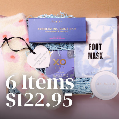 Gift box with fluffy bed socks, foot mask, XO tea, Scented candle reading "you are the best", exfoliating body bar and a ceramic keepsake ornament on a pink background.
