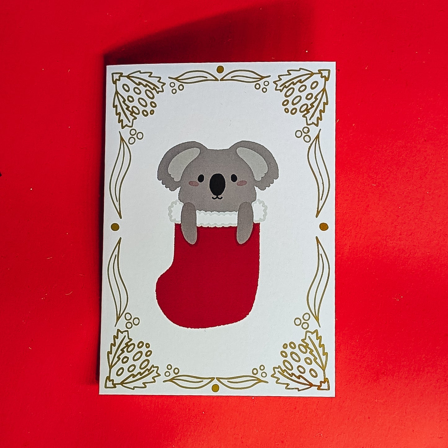Card with a koala in a red stocking on a white background, against a red border.
