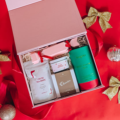 Pamper gift set with herbal tea, candle tin, chocolates, body pamper trio and a christmas bon bon on a red background with gold bows and christmas baubles.