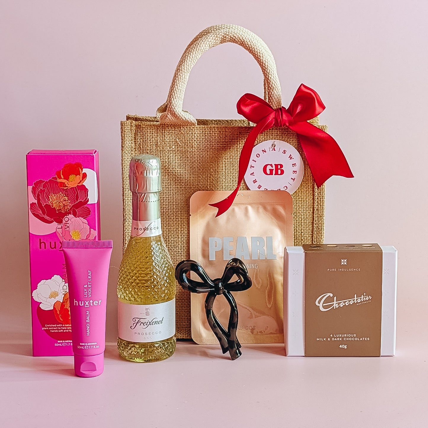 Gift set with a jute bag, wine bottle, chocolate, korean sheet mask and hand cream on a pink background.