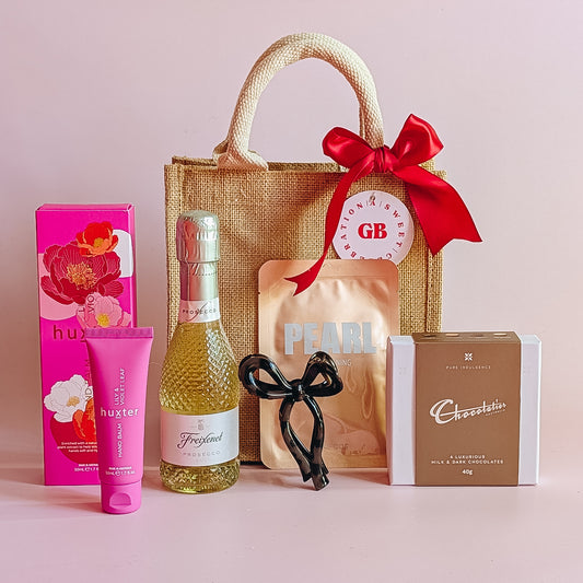 Gift set with a jute bag, wine bottle, chocolate, korean sheet mask and hand cream on a pink background.