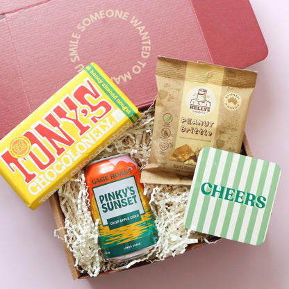 Congratulations gift for him overhead flatlay showing Gage Roads Pinky's Sunset cider, Tony's Chocolonely, Kelly's Peanut Brittle and Cheers drink coaster in open red gift box
