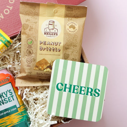 Congratulations gift for him close up showing Kelly's Peanut Brittle and Cheers stripey drink coaster with Gage Roads cider on pink background